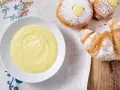 Una ciotola di soffice crema pasticcera senza uova accanto a cornetti e bomboloni spolverati di zucchero a velo.