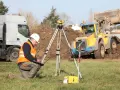 Geodeta w kamizelce odblaskowej nadzoruje odwierty geologiczne pod budowę domu. W tle koparka i ciężarówka.
