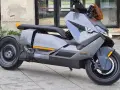 Szary skuter elektryczny BMW CE 04 z pomarańczowymi akcentami stoi na bruku przed budynkiem z dużymi oknami.