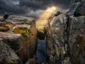 Enorm sten balanserar mellan klippor i Norge. Dramatiskt ljus bryter igenom molnen över en djup fjord.