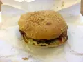 Burger z sezamem na białym papierze, widoczne składniki: mięso, sos, warzywa.