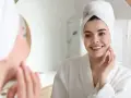 Uśmiechnięta kobieta w szlafroku i ręczniku na głowie dotyka policzka, zastanawiając się, jaki peeling do cery dojrzałej wybrać.