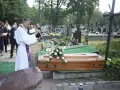 Wojciech Magnuski żegna zmarłego na cmentarzu. Ksiądz w białej szacie czyta z księgi przy trumnie ozdobionej białymi różami.