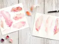 Aquarelles de plumes roses et orangées sur papier, avec des godets de peinture acrylique et un pinceau.
