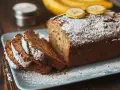 Wilgotne ciasto bananowe, posypane cukrem pudrem, z plasterkami banana na wierzchu, prezentuje się apetycznie na talerzu.