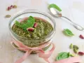 Un vasetto di pesto di pistacchi, decorato con un nastro rosa e guarnito con foglie di basilico e pistacchi interi, pronto per essere gustato.