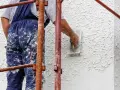Wykonawca w niebieskich, ubrudzonych farbą ogrodniczkach nakłada tynk baranek na elewację.