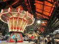 Drehende Kettenkarussells und bunte Lichter auf einem historischen Jahrmarkt in einer alten Industriehalle.