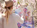 En kvinna med flätat blont hår målar en abstrakt tavla med olika målartekniker under ett körsbärsträd i blom.