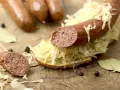 Lungenwurst zubereiten: Eine Scheibe Brot mit Sauerkraut und Lungenwurst belegt, daneben weitere Würste, Gewürze und Lorbeerblätter auf Holzuntergrund.