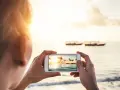 Une personne prend une photo d'un coucher de soleil sur la mer avec des bateaux au loin. Une belle photo à faire à la plage.
