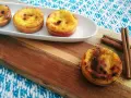 Vier goldbraune, karamellisierte Pastéis de Nata auf einem weißen Teller, daneben Zimtstangen auf einem Holzbrett.