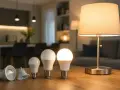 Różne rodzaje żarówek LED na drewnianym stole, od reflektorowych po kuliste, obok zapalonej lampki.