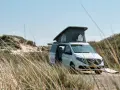 Vit el campervan med uppfällt tak står parkerad vid en sanddyn. En surfbräda ligger bredvid.