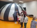 Dzieci i opiekunki wchodzą do dmuchanej kopuły, która przypomina planetarium. To miejsce, gdzie można oglądać filmy z przesłaniem, odkrywając tajemnice wszechświata.