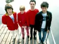 Die Band Talking Heads steht auf einem Holzsteg. David Byrne trägt ein rotes Hemd, Tina Weymouth ein pinkes Outfit.