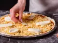 Dłoń układa kawałki sera na pizzy, która jest już posypana startym żółtym serem.