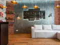 Nowoczesny salon w stylu loft: szara kanapa, ceglana ściana, panel z nocnym widokiem miasta i złote lampy.