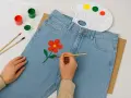 En person målar en röd blomma med gul mitt på ett par ljusblå jeans. Penslar, färger och en palett ligger bredvid.