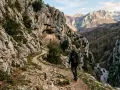 Un escursionista percorre un sentiero roccioso verso un eremo incastonato nella roccia, un'avventura nei cammini in Abruzzo.