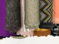 Rouleaux de tissus aux textures variées : noir uni, violet, motifs complexes, rose pâle, beige chiné, zigzags multicolores, pêche, et noir et blanc. Une matière mesh se distingue par sa finesse.