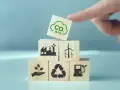 Klocki symbolizujące zielony ład: fabryka, wiatraki, sadzonka, recykling, paliwo. Na górze kostka z chmurką CO2 i strzałkami w dół.