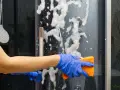 Main bleue, éponge orange pour nettoyer paroi douche. Mousse et bulles sur la paroi vitrée.