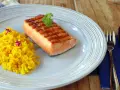 Un delicioso salmón a la plancha con marcas de parrilla, servido con arroz amarillo y decorado con perejil y flores moradas.