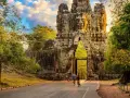 Two cyclists approach a massive stone gateway, one of the ancient Cambodia temples, bathed in golden sunlight.