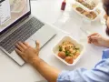 Mężczyzna pracuje przy laptopie i je dietetyczny posiłek z pudełka.