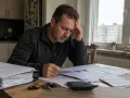 Zmartwiony mężczyzna analizuje dokumenty, szukając odpowiedzi, jak zachować mieszkanie przy upadłości. Na stole leżą klucze i kalkulator.