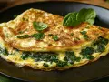 Złocisty omlet ze szpinakiem, udekorowany listkami bazylii i natki, prezentuje się apetycznie na ciemnym talerzu.