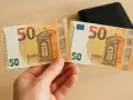 Un billet de 50 euros 2002, coupé en deux, est tenu dans une main. Un portefeuille noir est visible en arrière-plan.