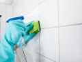 Manos con guantes azules rociando limpiador y frotando juntas de azulejos blancos con una esponja verde para blanquear juntas azulejos.