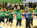Krystian Woźniak i jego drużyna świętują zwycięstwo. Zawodnicy w zielonych strojach przytulają się na boisku, a na trybunach widać kibiców.