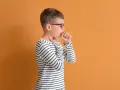 Chłopiec w okularach ma **szczekający kaszel u dziecka**. Dotyka gardła, czując dyskomfort.