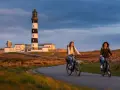 Deux femmes font du vélo sur une route sinueuse à Ouessant, avec un phare emblématique en arrière-plan. Une belle façon de visiter Ouessant à pied, ou plutôt à vélo !