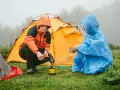 Zwei Camper in Regenponchos bereiten sich auf das Zelten bei Regen vor. Einer kocht auf einem Campingkocher, der andere hält eine Tasse.