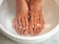 A relaxing home remedy foot soak in a white basin. Feet with french manicures and one toe nail decorated with a black and white design are submerged in clear water.