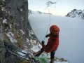 Wspinaczka górska w chmurach. Alpinista w pomarańczowym kasku i kurtce przygotowuje się do zjazdu na linie, otoczony zimowym krajobrazem i mgłą.