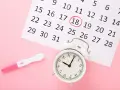 Test ciążowy i kalendarz z zaznaczonym dniem 18. Zegar obok sugeruje, że czas na badania prenatalne kiedyś nadejdzie.
