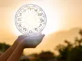 Dłonie trzymają koło zodiaku, gdzie widoczny jest symbol Wagi. Słońce przebija się przez niebo, tworząc magiczną aurę.