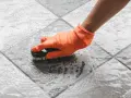 Mano con guanto arancione che spazzola un pavimento piastrellato bagnato, mostrando come togliere ruggine dal pavimento con schiuma.