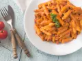 Piatto di penne alla vodka cremose, guarnite con prezzemolo, su un tavolo di legno azzurro. Accanto, pomodorini freschi e posate rustiche.
