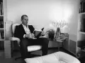 Un homme lit un livre dans un fauteuil, entouré de livres. La lumière d'une lampe éclaire la scène. ae-l af-l.