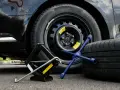 Kit per cambiare una ruota: cric, chiave a stella e ruota di scorta accanto a un'auto nera.