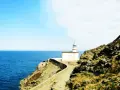 Un faro blanco se alza sobre un acantilado rocoso, vigilando el mar azul. Un camino serpentea hacia él, invitando a explorar los **faros Costa Brava**.