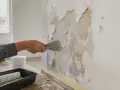 Ręka zeskrobuje łuszczącą się farbę ze ściany, przygotowując ją do gruntowania.