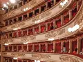 Widownia La Scali, pełna widzów w lożach, oczekuje na rozpoczęcie spektaklu. Bogaty repertuar teatru przyciąga miłośników sztuki.