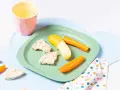 Baby-gerechte Snacks: Reiswaffeln, Karottensticks und eine halbe Banane auf einem grünen Teller.